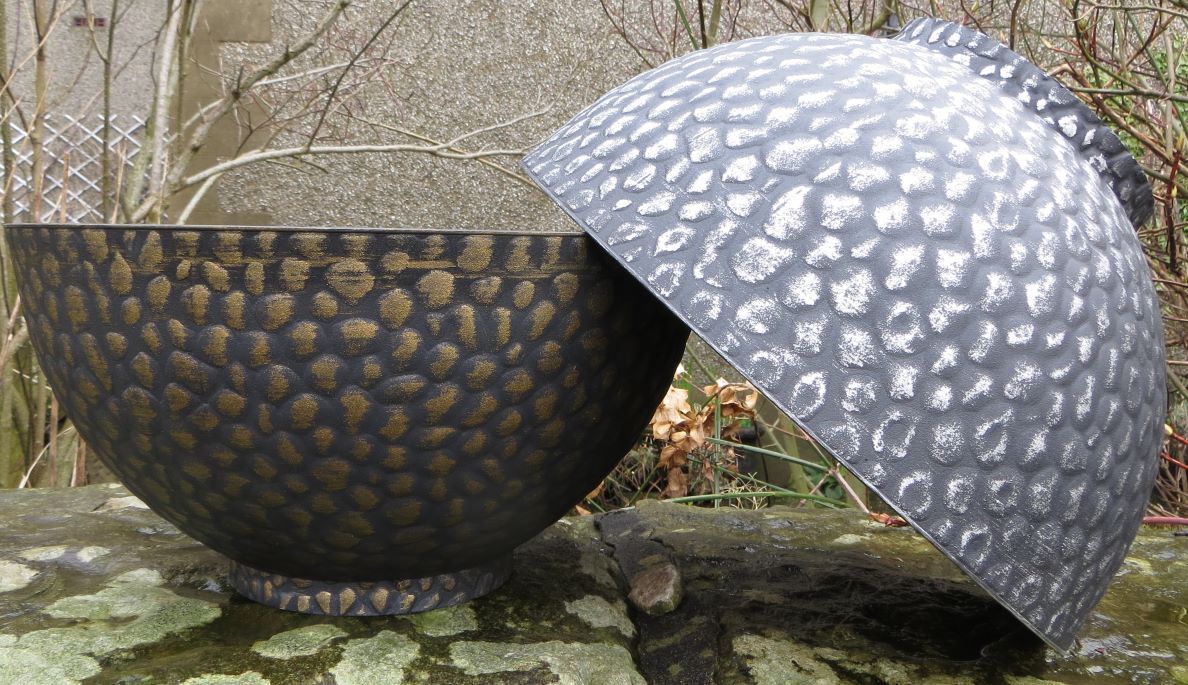 Two textured decorative plant bowls in metallic finishes, one upright in black and bronze, the other tilted in silver and grey, displayed outdoors on a mossy stone surface.